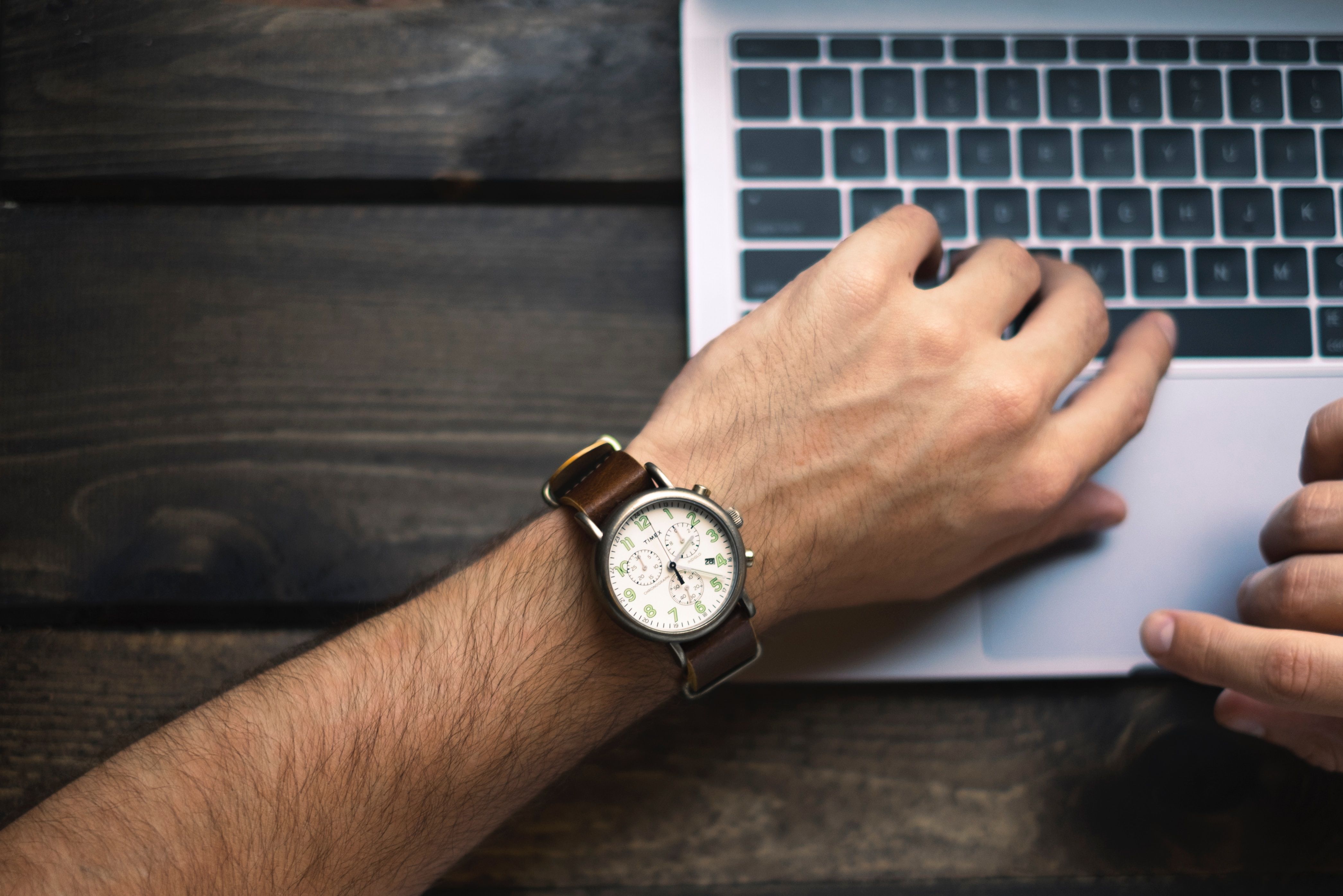 A man's wrist with a traditional watch with a laptop in the background