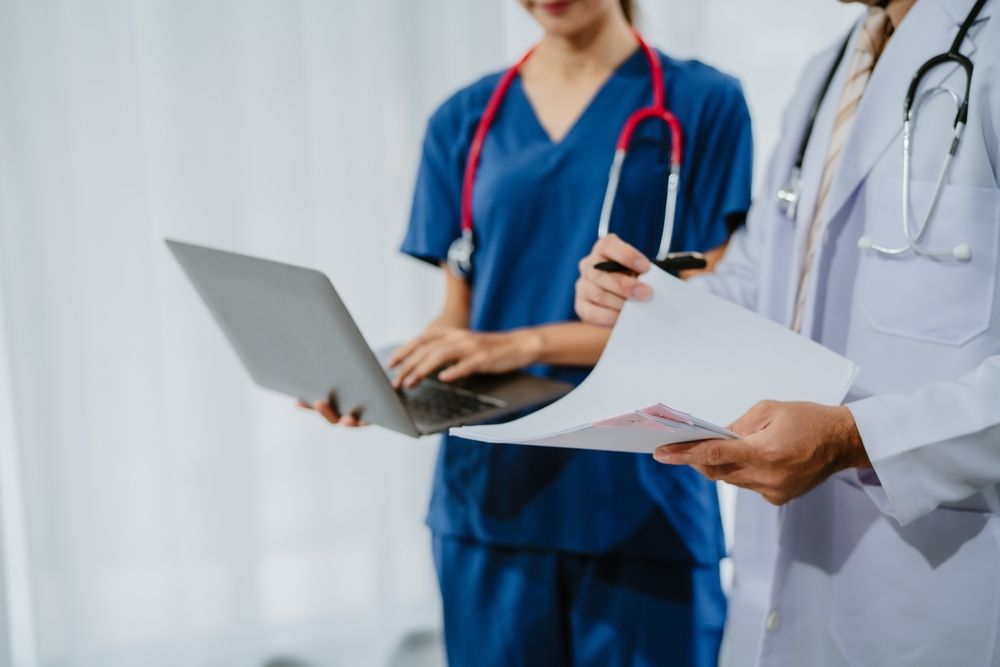 a male doctor holding paper patient records, which female medical nurse holding laptop.
